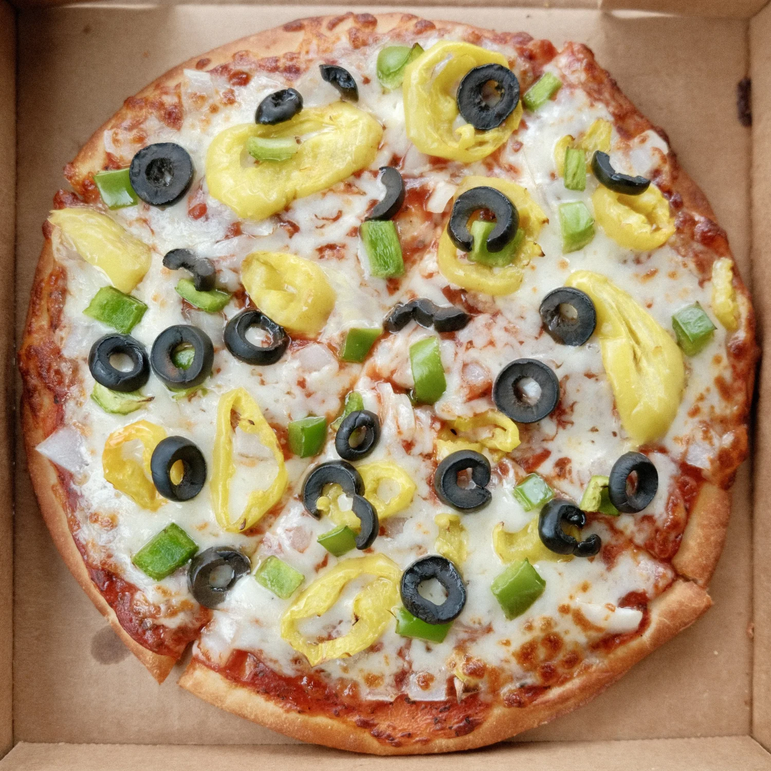 A whole Angilo's pizza in an open carryout box, topped with banana peppers, black olives, green peppers and onion.